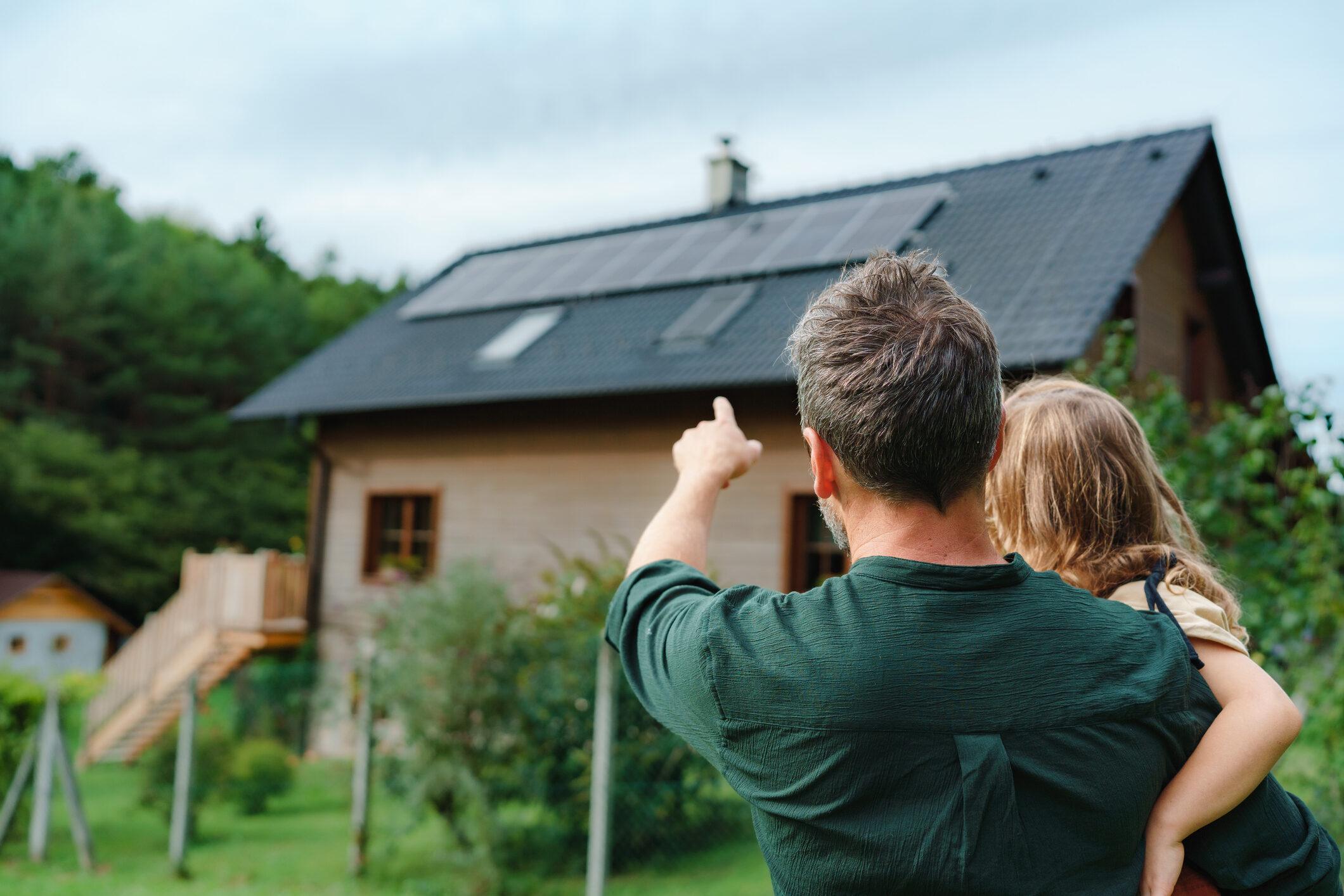 Rear view of dad holding her little girl in arms and showing at their house with solar panels. Alternative energy, saving resources and sustainable lifestyle concept.